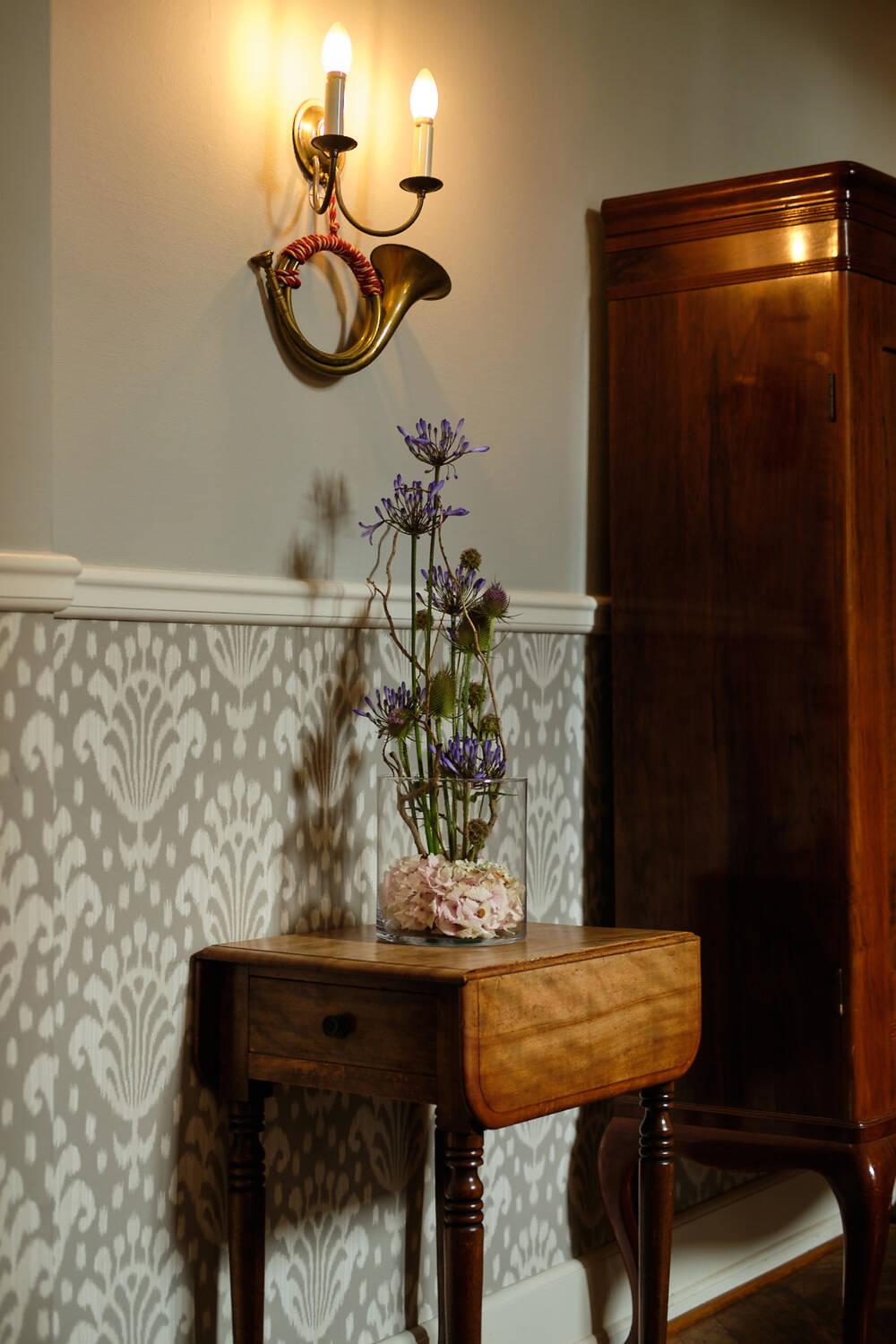 Ein kleiner Holztisch mit einer Blumenvase unter einer Wandleuchte in Form eines Horns.