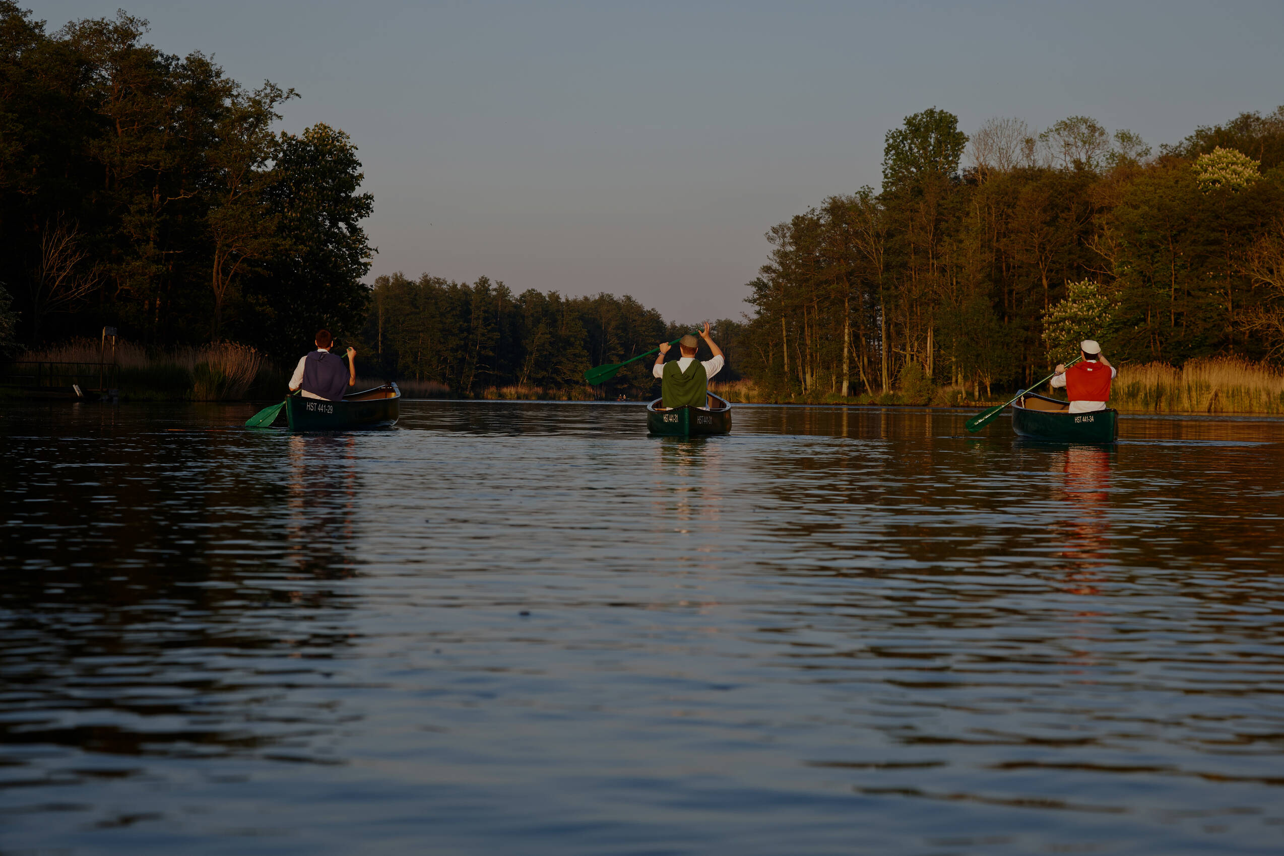 Drei Personen paddeln in Kanus auf einem stillen Fluss, umgeben von Bäumen bei sanftem Abendlicht.