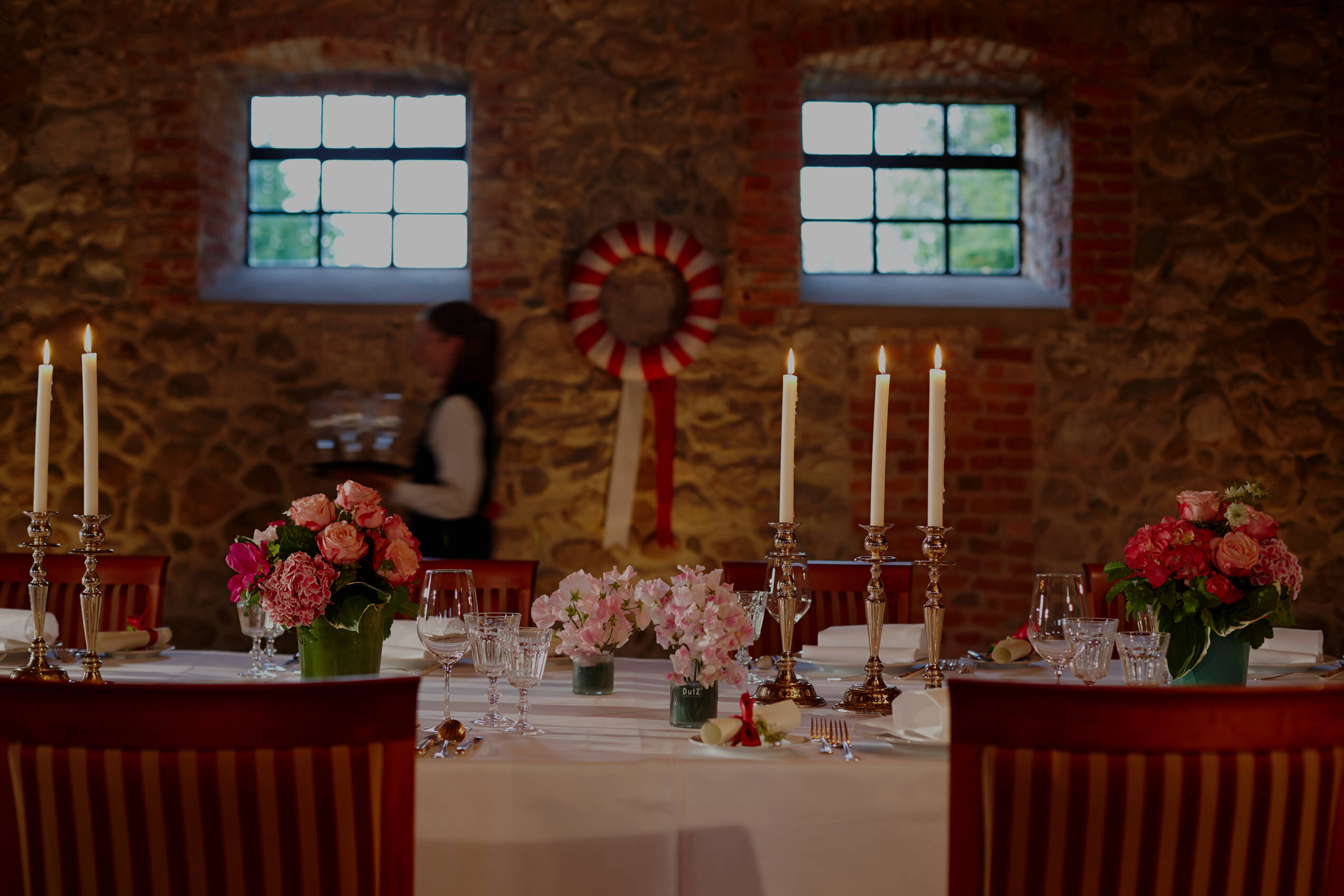 Ein festlich gedeckter Tisch mit Kerzenleuchtern, Blumenarrangements und Gläsern vor einer Steinwand mit zwei Fenstern.