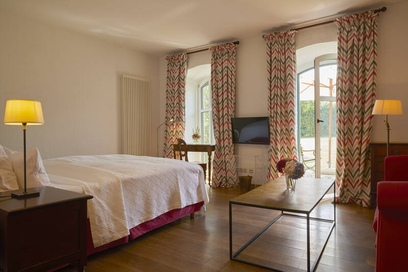 Ein großzügiges Hotelzimmer mit Doppelbett, farbigen Vorhängen, Schreibtisch und Zugang zur Terrasse mit Blick ins Grüne.