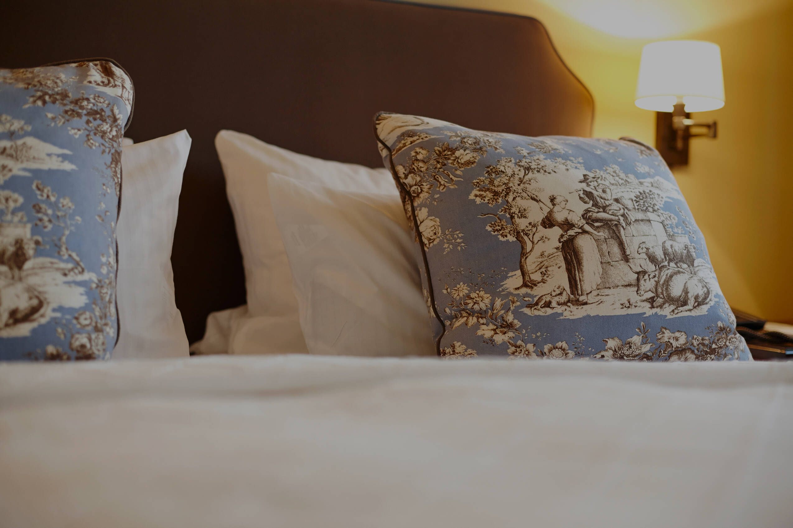 Nahaufnahme eines Hotelbetts mit weißen Laken und blauen Zierkissen mit klassischem Motiv unter warmem Licht.