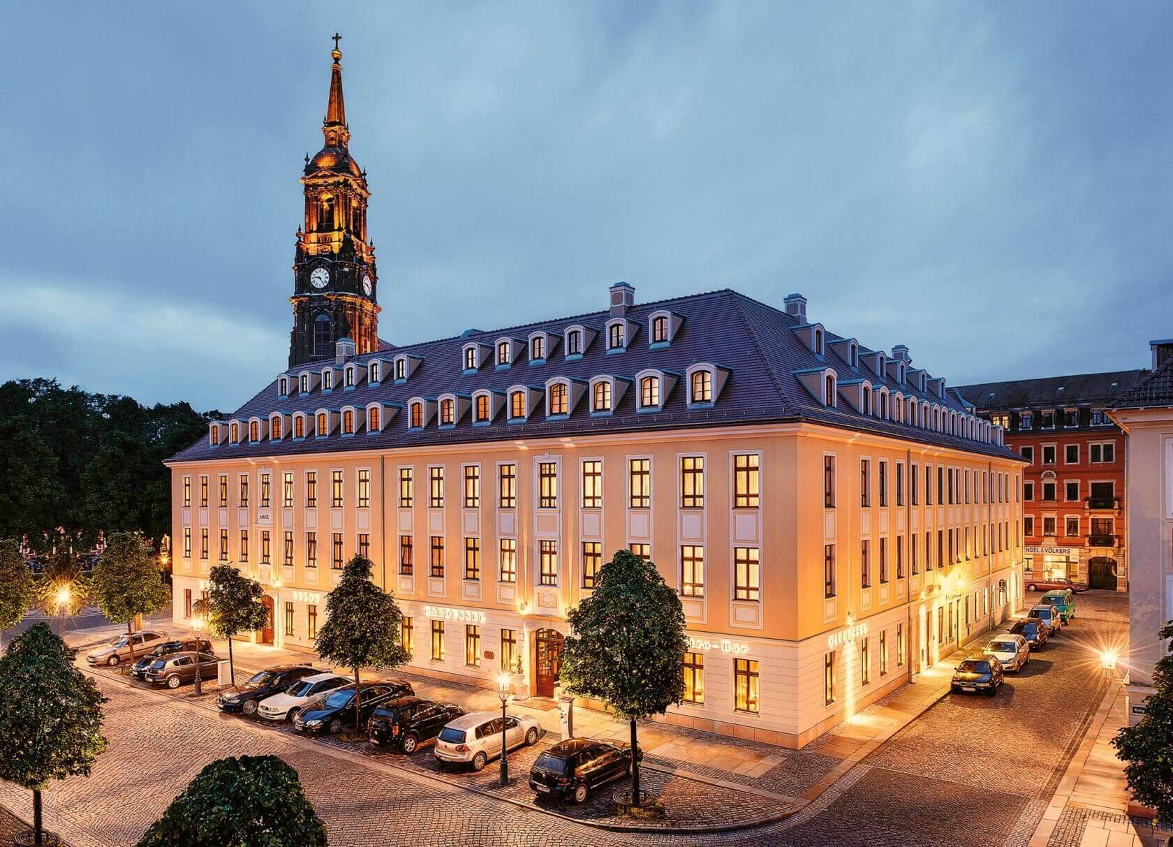 Ein beleuchtetes Stadthotel mit einer klassischen Fassade und Kirchturm im Hintergrund bei Abendstimmung.