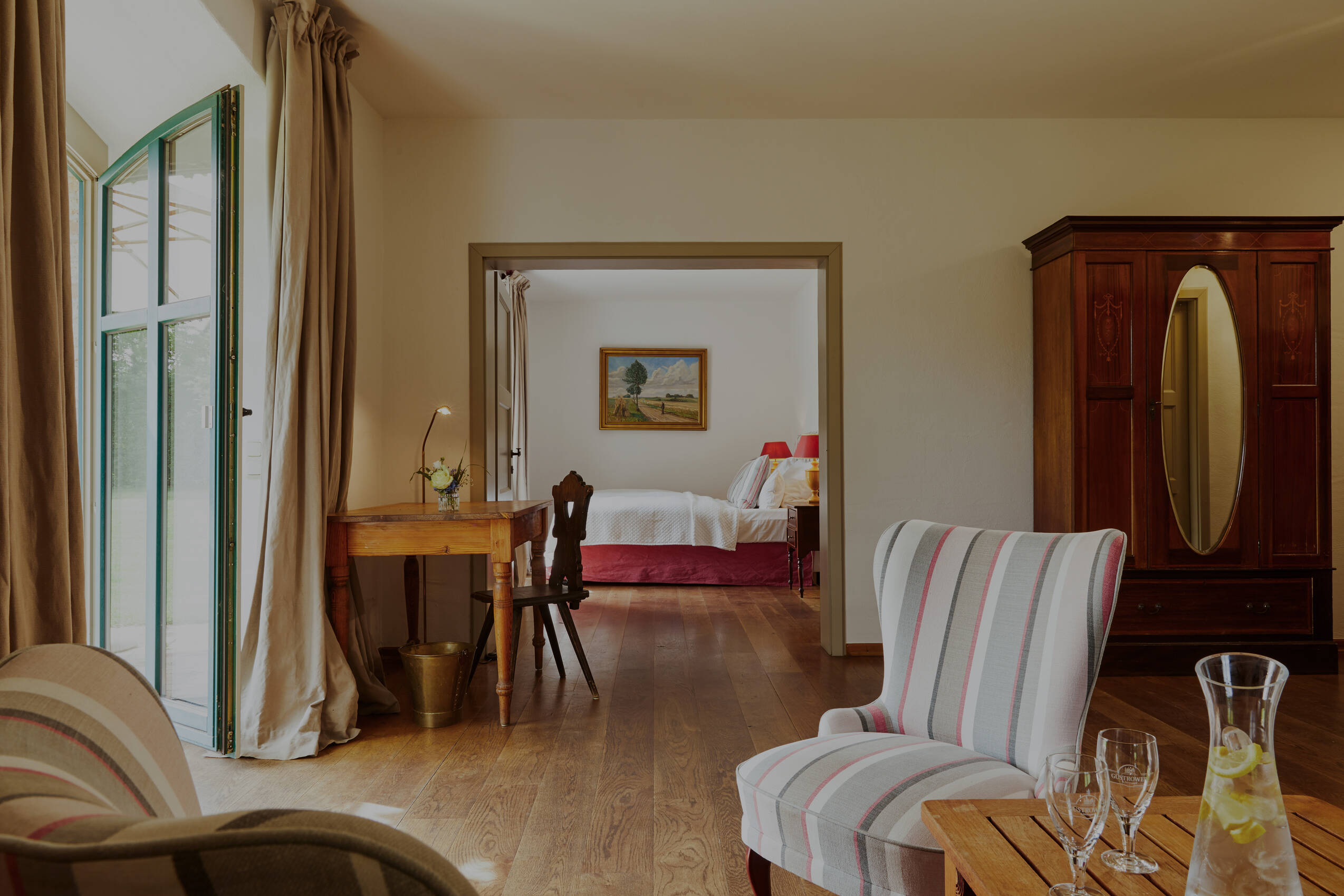 Eine helle Suite mit Sesseln, einem Holztisch und einer Kommode und Terrasse, Blick durch die Tür in ein Schlafzimmer mit einem Bett.