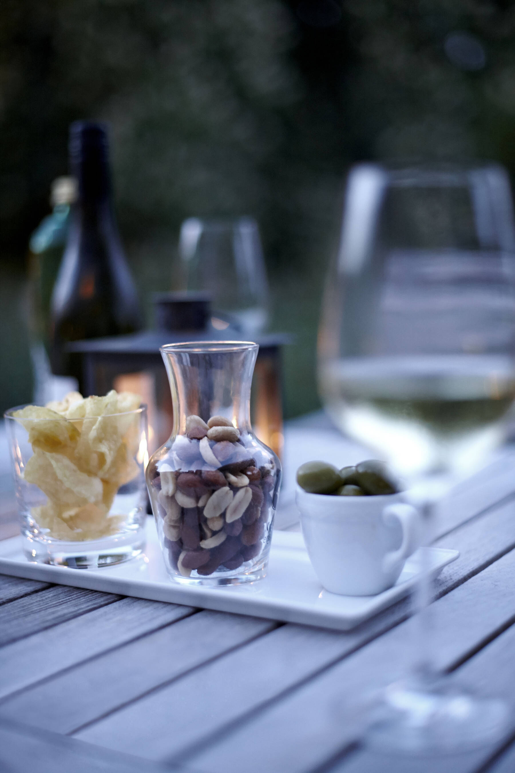 Ein Holztisch mit Wein, Oliven, Nüssen und Chips steht im sanften Abendlicht.