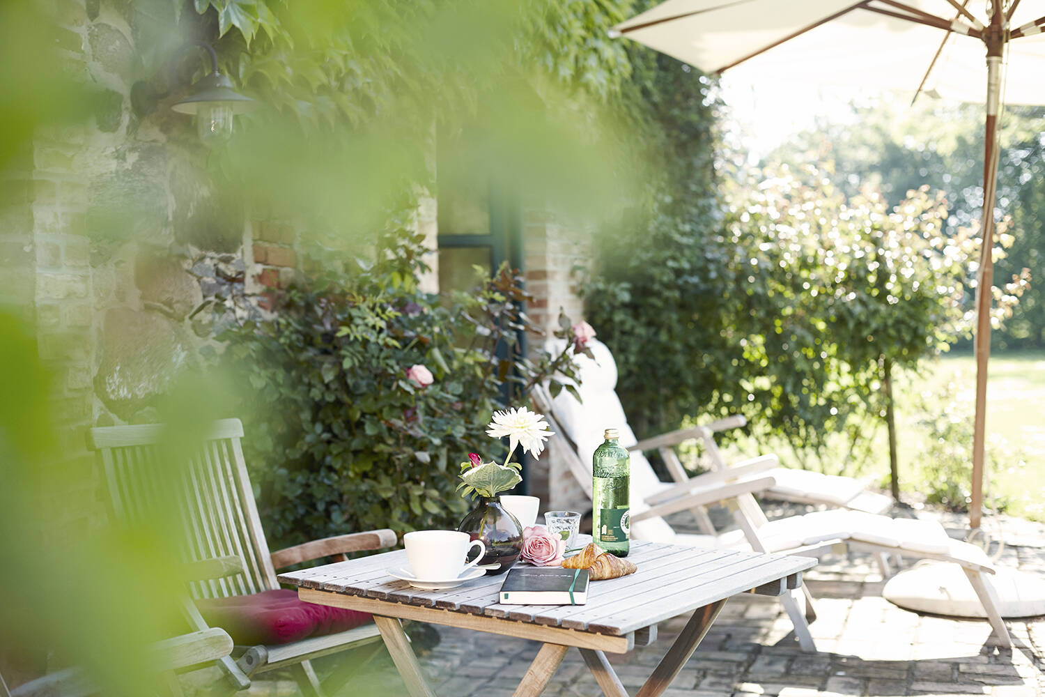 Ein Holztisch mit Kaffee, Croissant und Blumen steht auf einer sonnigen Terrasse mit Blick in den Garten.