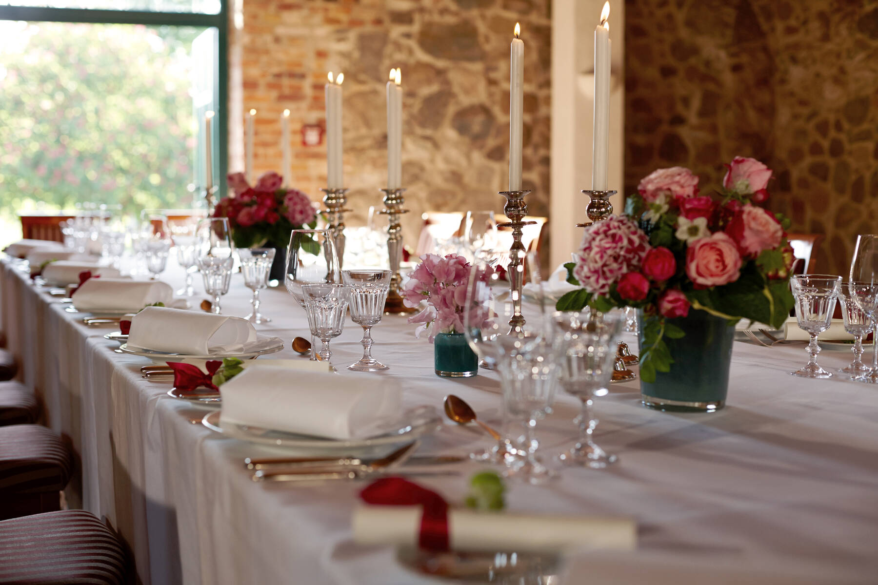 Ein festlich gedeckter Tisch mit weißen Kerzen, Kristallgläsern und Blumenarrangements in einem Raum mit einer Steinwand.