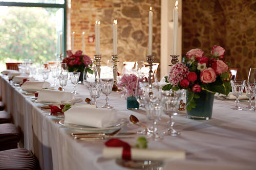 Ein festlich gedeckter Tisch mit weißen Kerzen, Kristallgläsern und Blumenarrangements in einem Raum mit einer Steinwand.