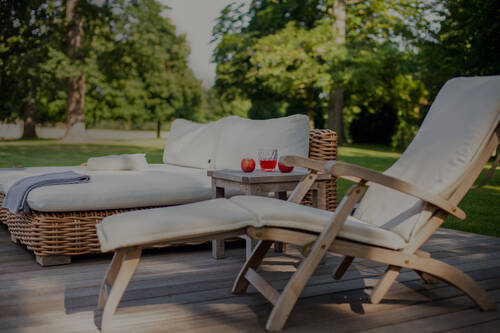 Ein Holzliegestuhl und eine Rattan Liege mit Kissen auf einer Holzterrasse; im Hintergrund Bäume und Rasen.