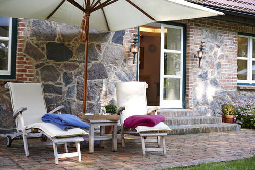 Zwei weiße Liegestühle mit bunten Decken stehen unter einem Sonnenschirm vor einem Steinhaus - dem SPA Bereich vom Gutshaus Stolpe.