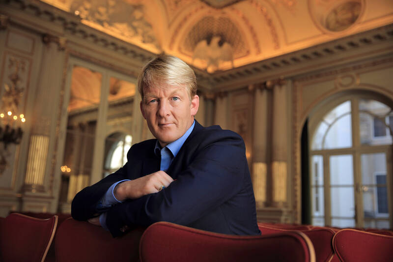 Ein Mann in einem dunklen Anzug sitzt mit verschränkten Armen auf einem roten Sofa in einem sehr prunkvollen Saal des Gutshaus Stolpe.