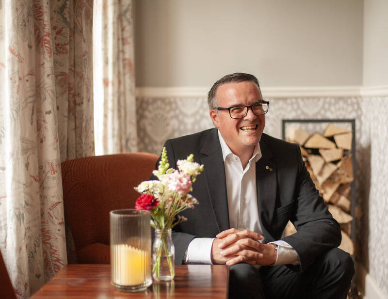 Ein Hotelmanager sitzt lächelnd in einem Sessel mit Blumen und Kerze auf dem Tisch, im Hintergrund gestapeltes Kaminholz.