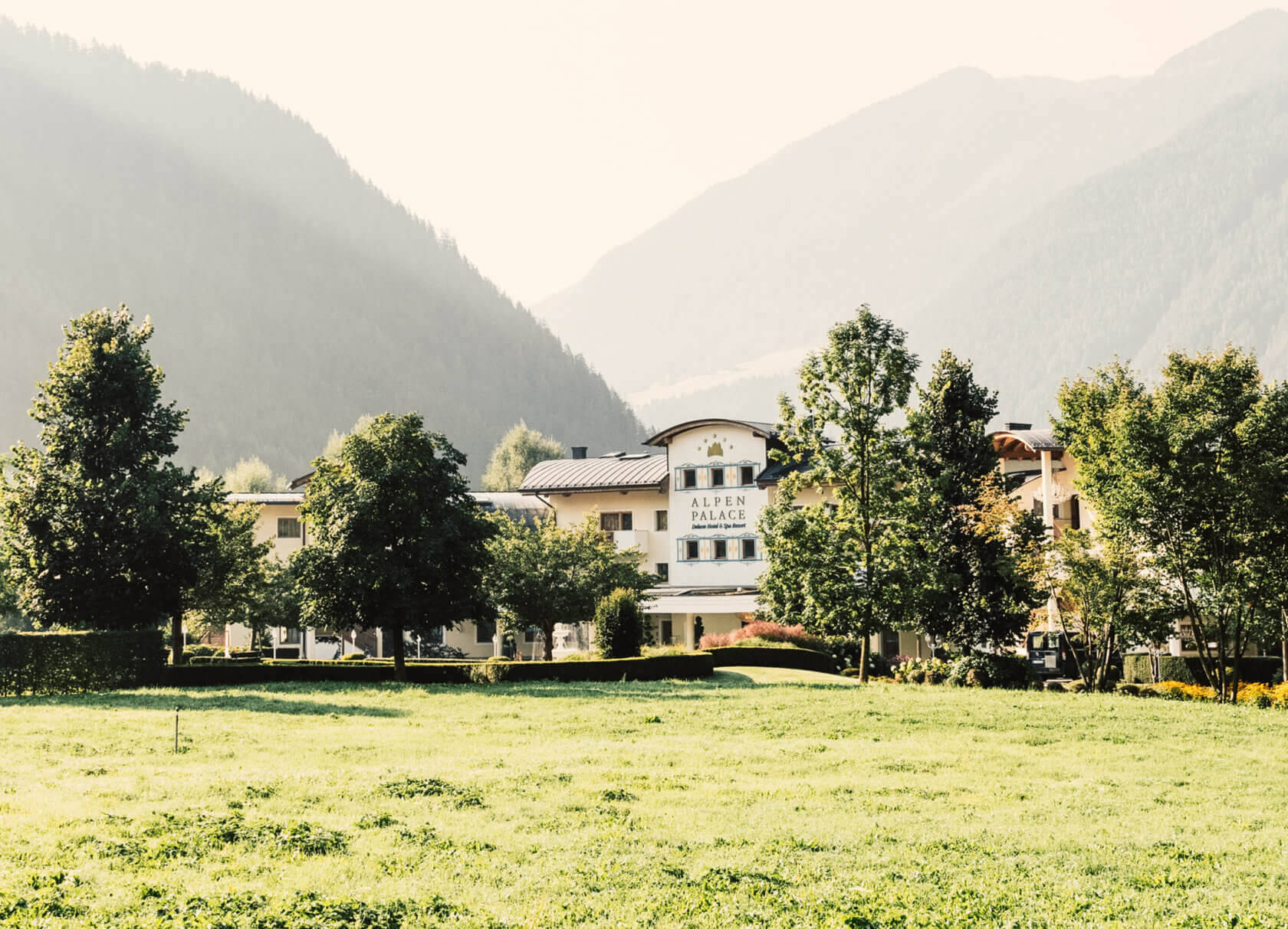 Ein Hotelgebäude in den Alpen vor bewaldeten Bergen und Wiesen im Vordergrund.
