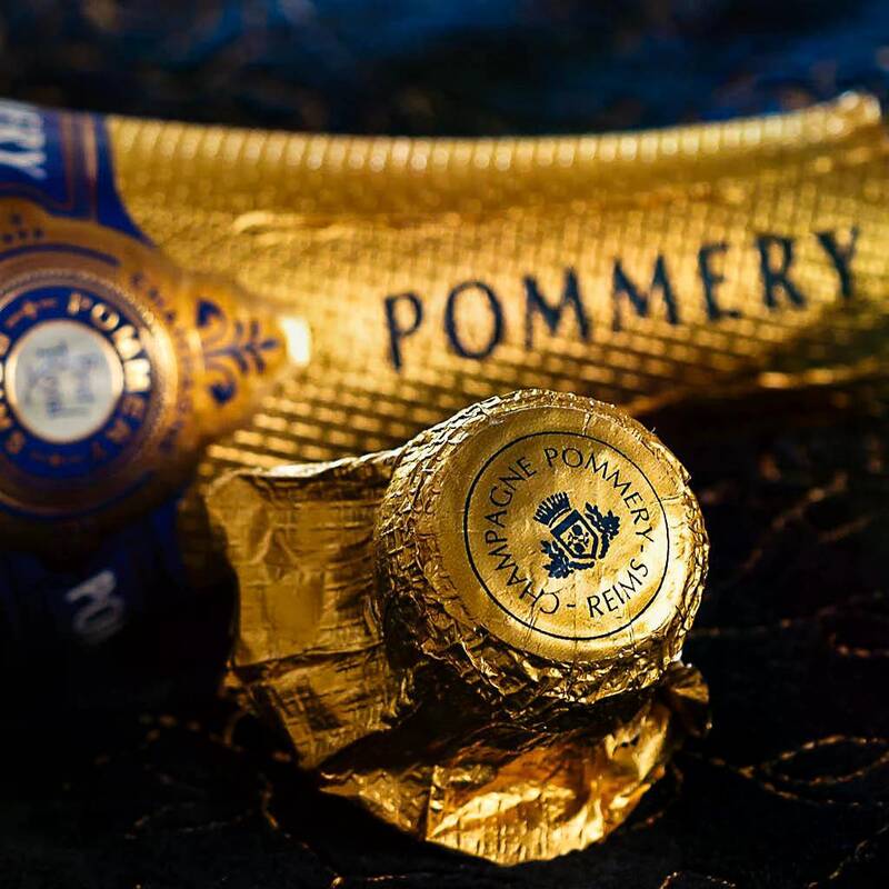 Eine goldene Champagnerflasche mit der Aufschrift "POMMERY" und einer goldenen Kapsel mit dem Text "CHAMPAGNE POMMERY REIMS" auf einem schwarzen Untergrund.