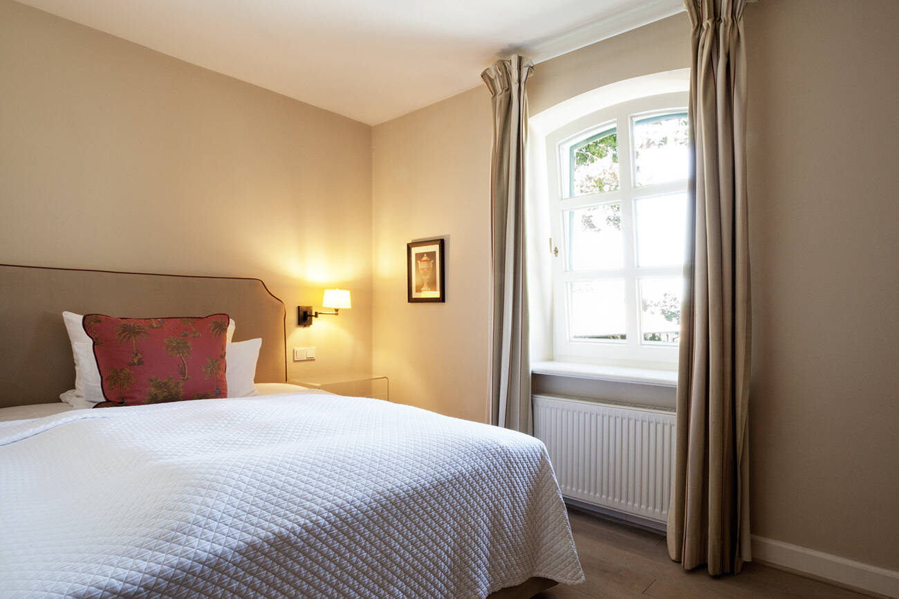 Ein Hotelzimmer mit einem Einzelbett, einer weißen Steppdecke, rotem Kissen, Wandlampe und einem Fenster mit hellbraunen Vorhängen.