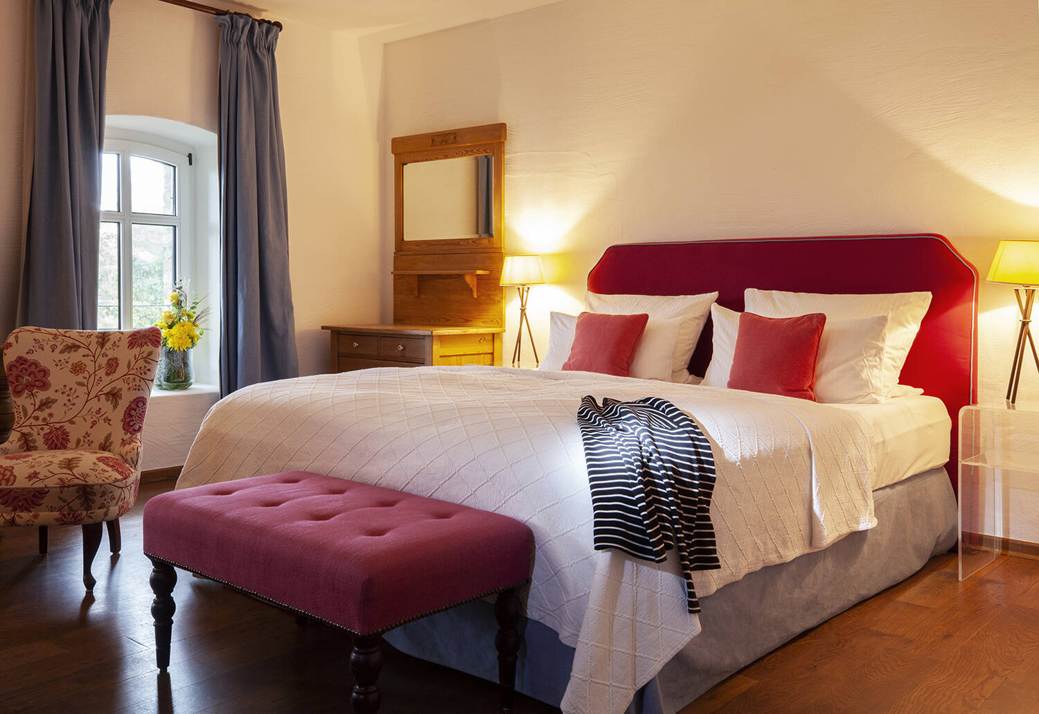 Ein Hotelzimmer mit einem großen Bett, Hocker, Sessel mit einem Blumenmuster, einem Fenster mit Vorhängen und zwei Lampen auf Nachttischen.