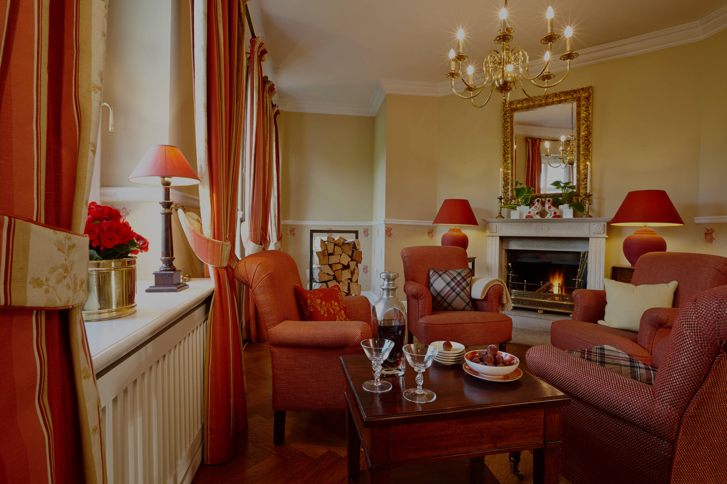 Ein gemütlicher Raum in Gutshaus Stolpe mit Kamin, zwei roten Sesseln, einem Sofa, Holztisch mit Gläsern und einer Obstschale, elegant orangenen Lampenschirmen.