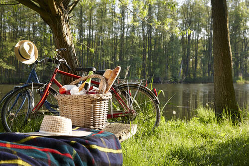Ein Fahrrad mit Weidenkorb voller Baguette und Getränken an einem See, daneben zwei Strohhüte auf einer Decke, im Hintergrund Bäume und Wasser.