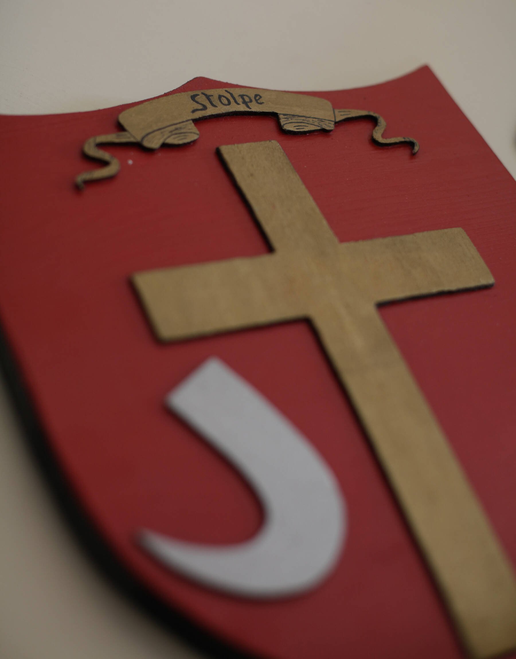 Ein rotes Wappenschild mit einem goldenen Kreuz, weißem Haken und einem Schriftband mit dem Wort "Stolpe".