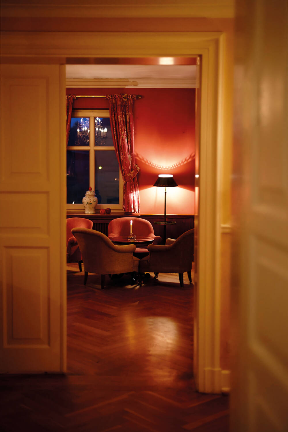 Ein einladender Blick in eine Lounge mit roten Wänden, Sesseln, Lampe und Kerzenschein durch eine geöffnete Tür.