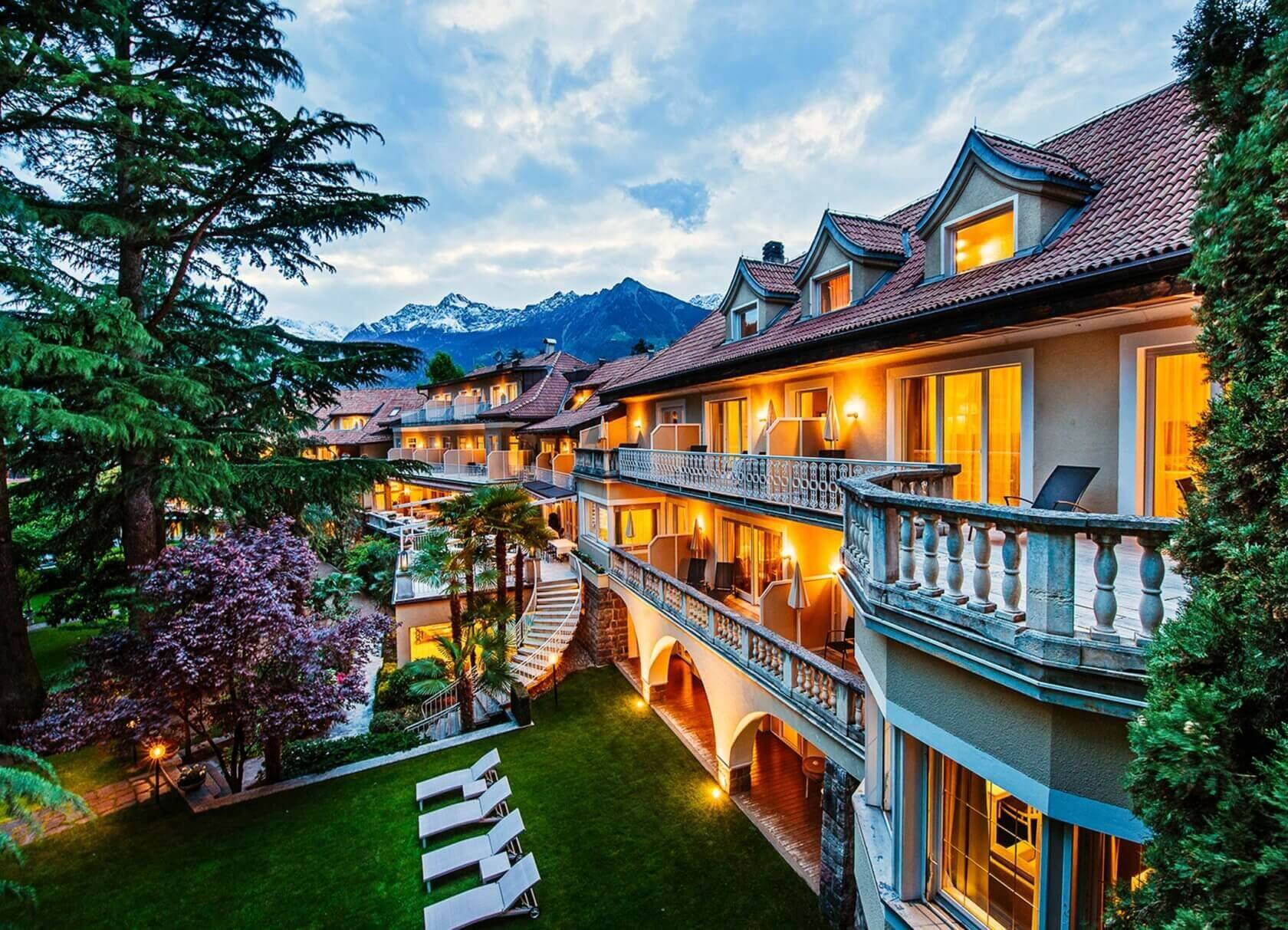 Ein mehrstöckiges Hotelgebäude mit beleuchteten Fenstern, Balkonen und Liegestühlen im Garten, umgeben von Bäumen und Bergen im Hintergrund.