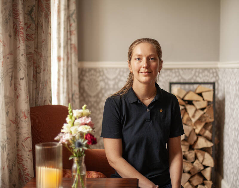 Eine junge Frau in einem dunklen Poloshirt sitzt in einem Sessel vor Kaminholz, einer Kerze und floralen Gardinen.