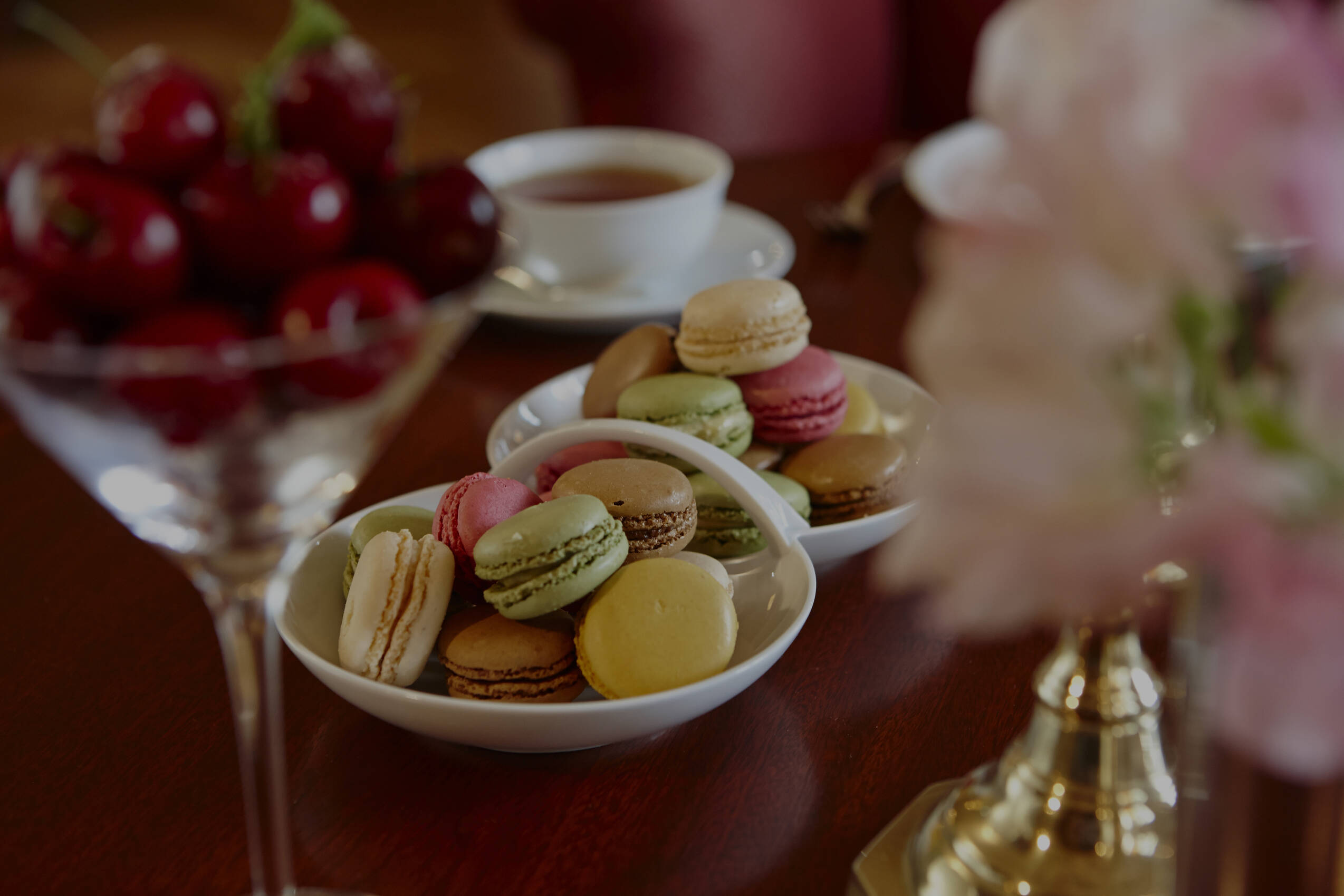 Bunte Macarons und frische Kirschen auf einem gedeckten Tisch mit Teetasse und Blumenschmuck.